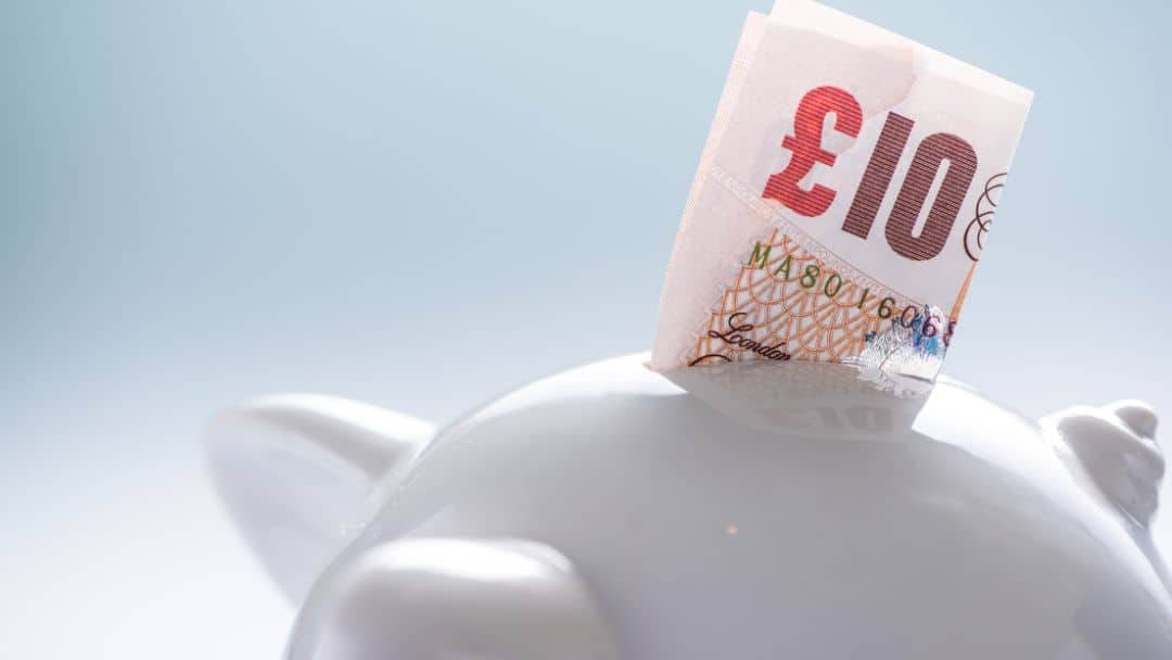 close up of a white piggy bank with a folded £10 note sticking out of the slot
