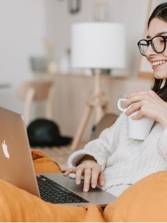 young woman with glasses relaxing at home on a sofa with a coffee browsing on a laptop