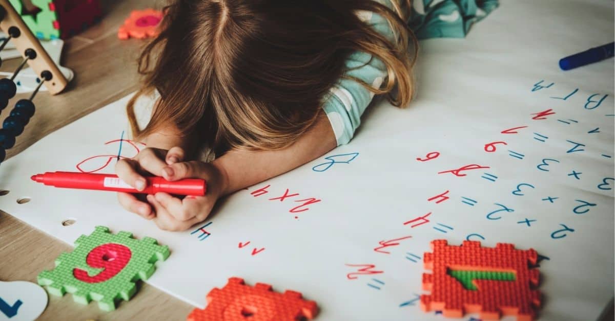 a little girl lies face down on the floor on top of a giant piece of paper full of times tables